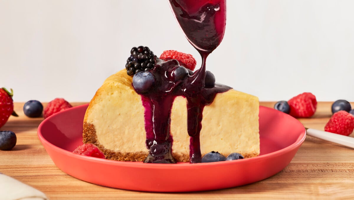 A slice of New York cheesecake on a plate with blueberry compote being drizzled on top.