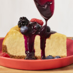 A slice of New York cheesecake on a plate with blueberry compote being drizzled on top.