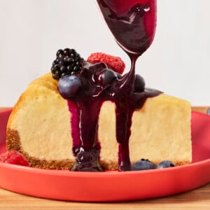 A slice of New York cheesecake on a plate with blueberry compote being drizzled on top.