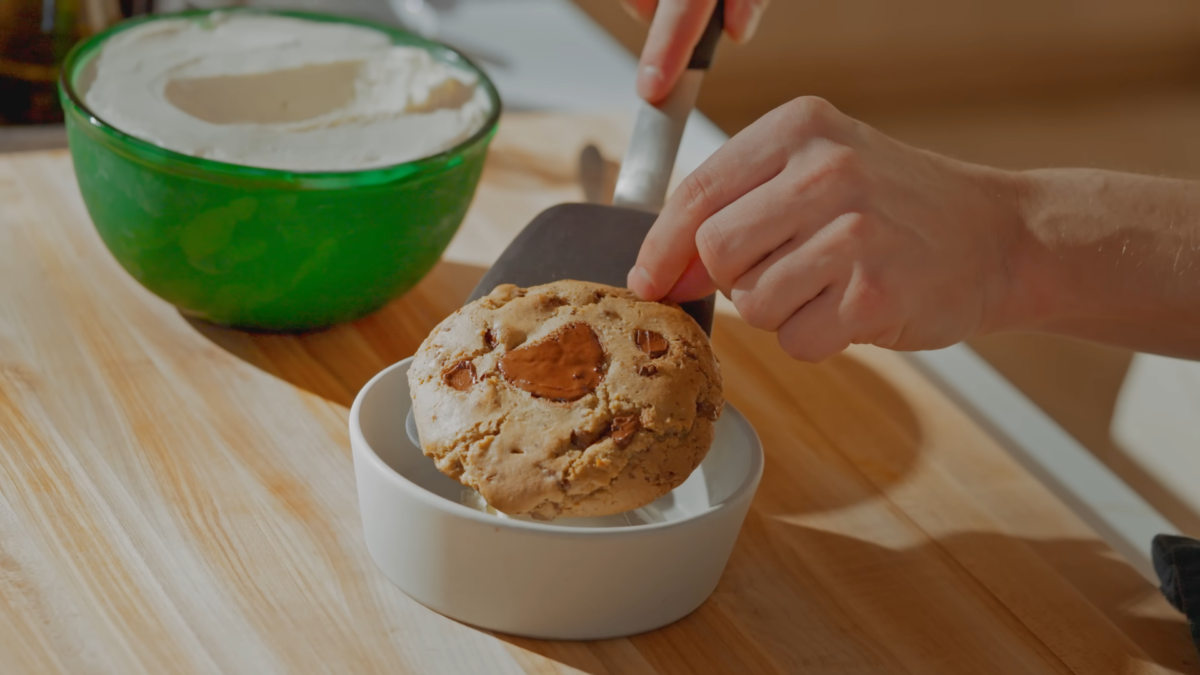 A chocolate chip cookie being laid on top of a scoop of ice cream in a bowl.