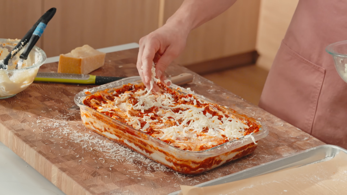 A male chef layering homemade lasagna in a glass baking dish.