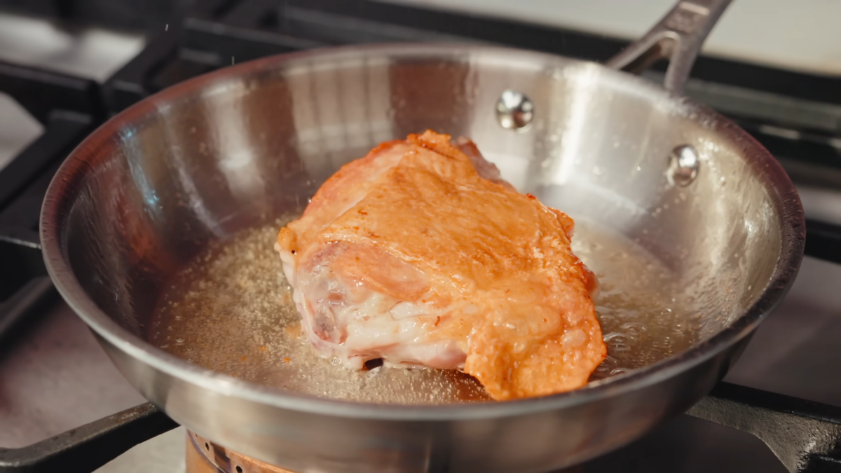 A chicken thigh frying in a metal pan.