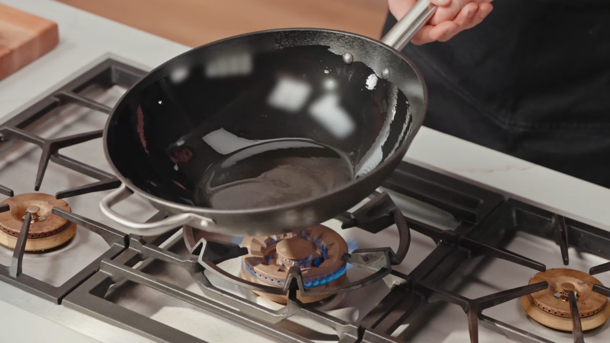A man holding a wok coated in oil over a stove.