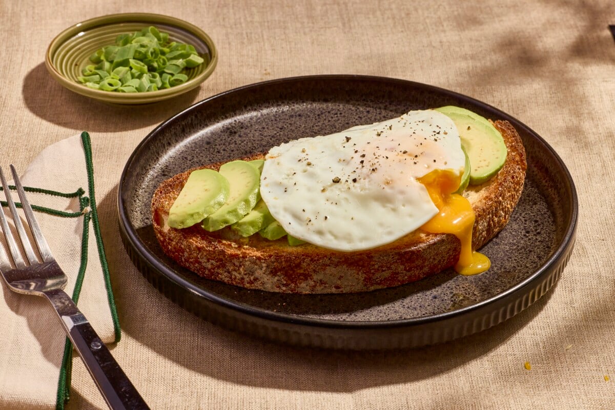 A slice of avocado toast on a plate topped with a sunny side up egg.