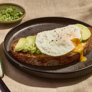 A slice of avocado toast on a plate topped with a sunny side up egg.