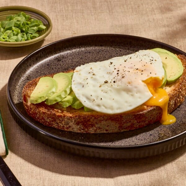 A slice of avocado toast on a plate topped with a sunny side up egg.
