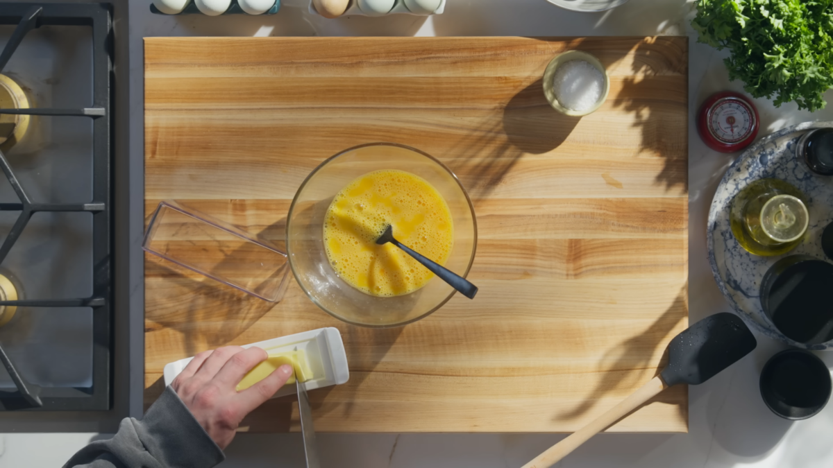 A glass bowl of whisked eggs on a wooden cutting board.
