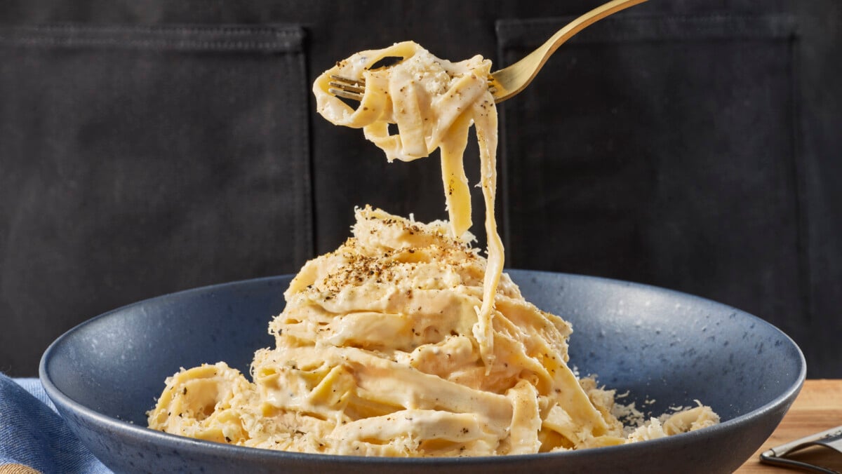 A fork holding a bite of fettuccine Alfredo above a bowl.