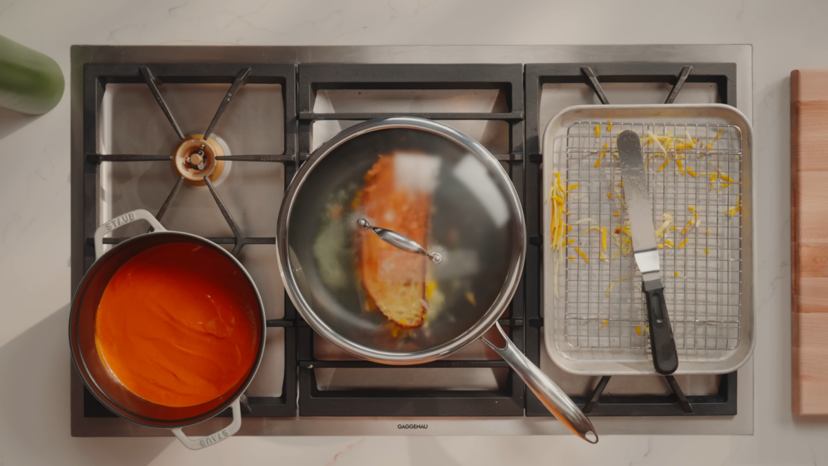 A stovetop with a pot of tomato soup, a covered pan with a grilled cheese sandwich, and a baking sheet.