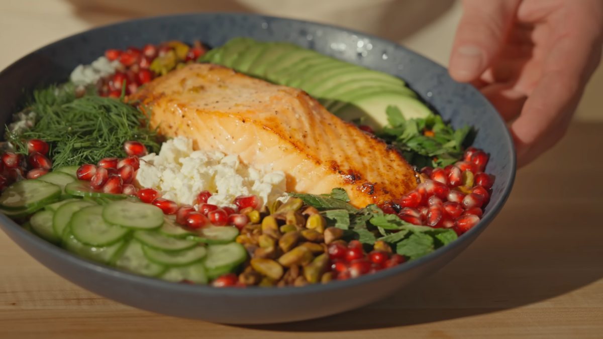 A grain bowl with salmon, pomegranate seeds, avocado slices, crumbled cheese, and herbs.