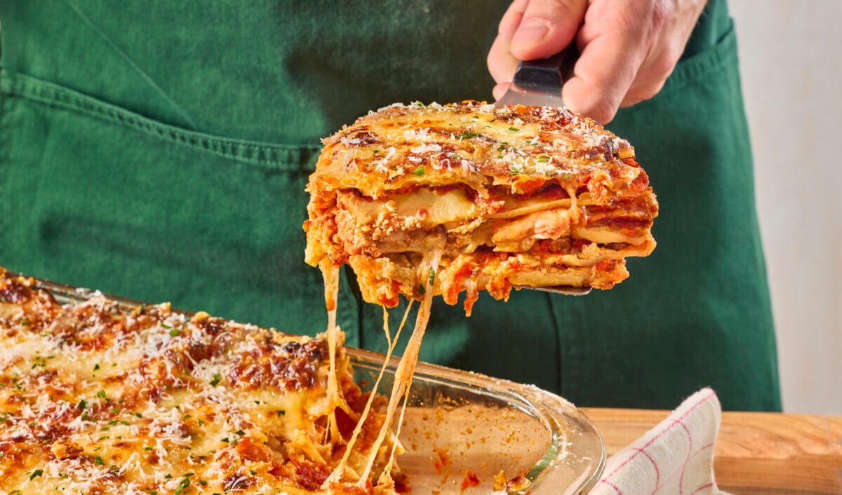 A male chef scooping gooey stringy lasagna out of a pan.