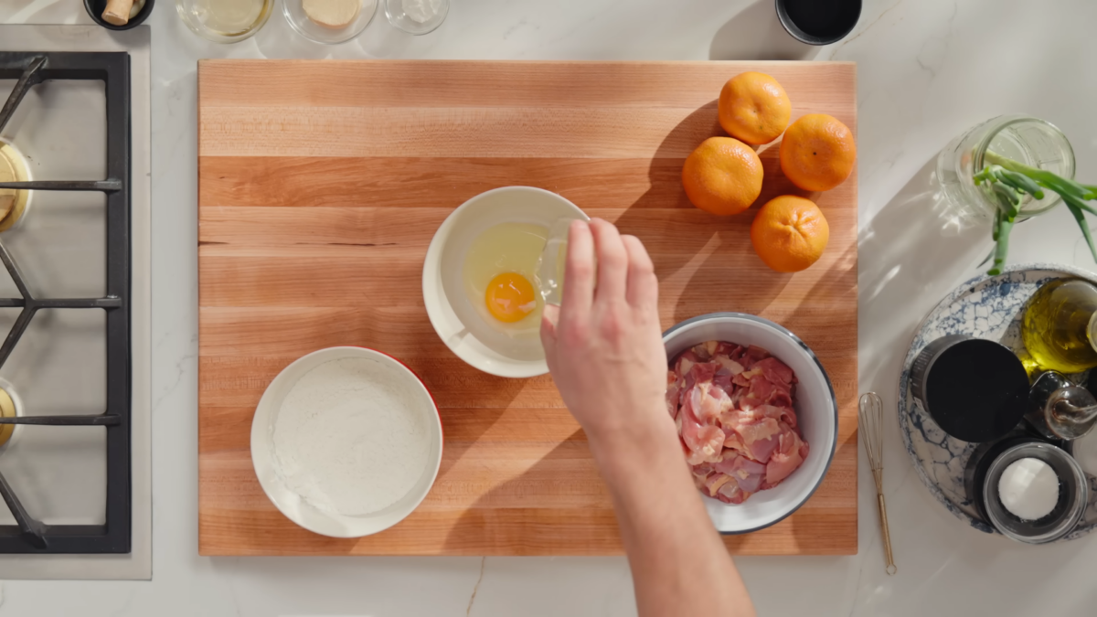 a cutting board with a bowl of flour, bowl of diced chicken, and a bowl with a raw egg.