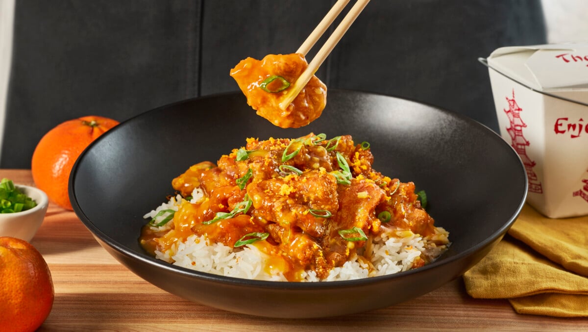 Chopsticks holding a piece of orange chicken over a bowl of orange chicken and rice.