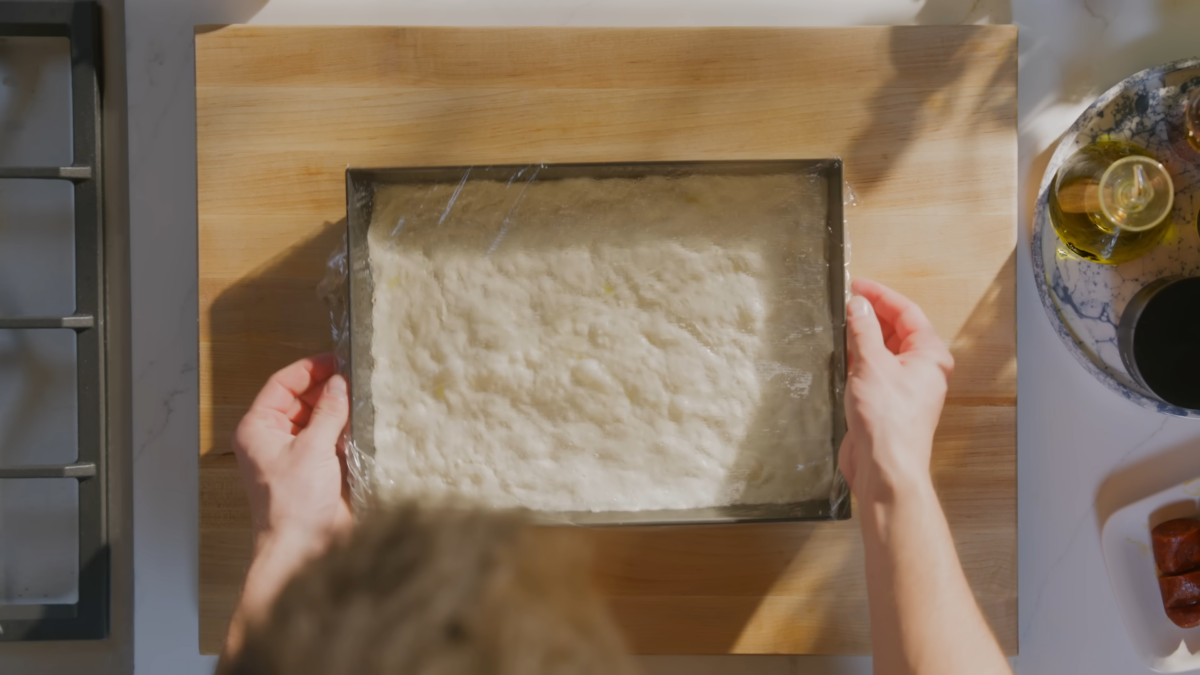 A baking dish with pizza dough covered with plastic wrap.