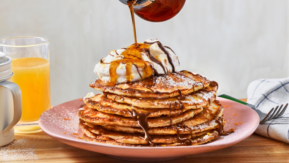 A stack of buttermilk pancakes with whipped cream with maple syrup being poured on top. 