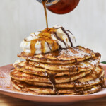 A stack of buttermilk pancakes with whipped cream with maple syrup being poured on top.