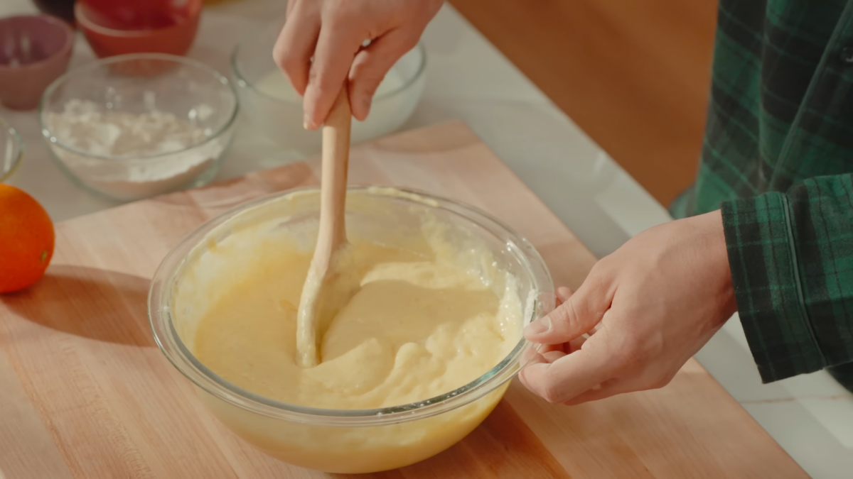 A man mixing pancake batter in a galss bowl with a wooden spoon.