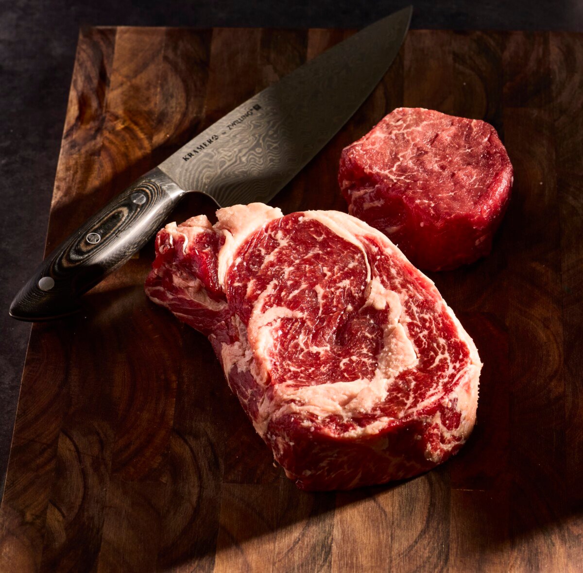 A raw ribeye steak and raw filet mignon on a cutting board with a knife.