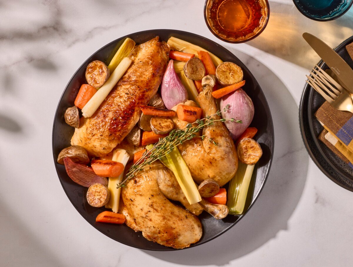 A rotisserie style whole roasted chicken on a plate with potatoes, carrots, shallots, and leeks.