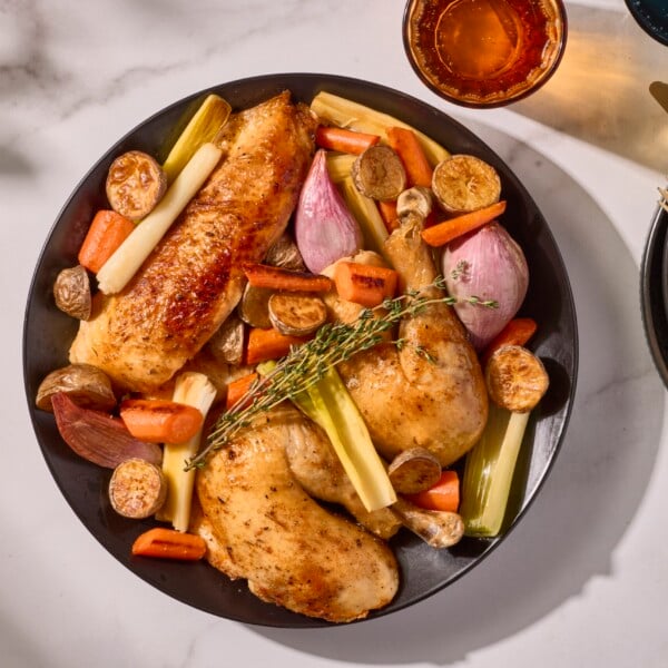 A rotisserie style whole roasted chicken on a plate with potatoes, carrots, shallots, and leeks.