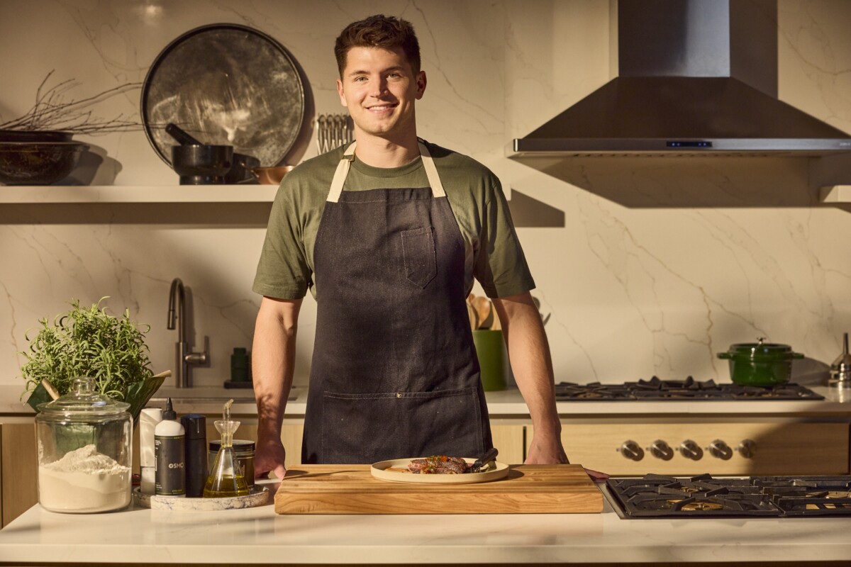 A male chef standing at a counter behind a plate of steak on a counter.