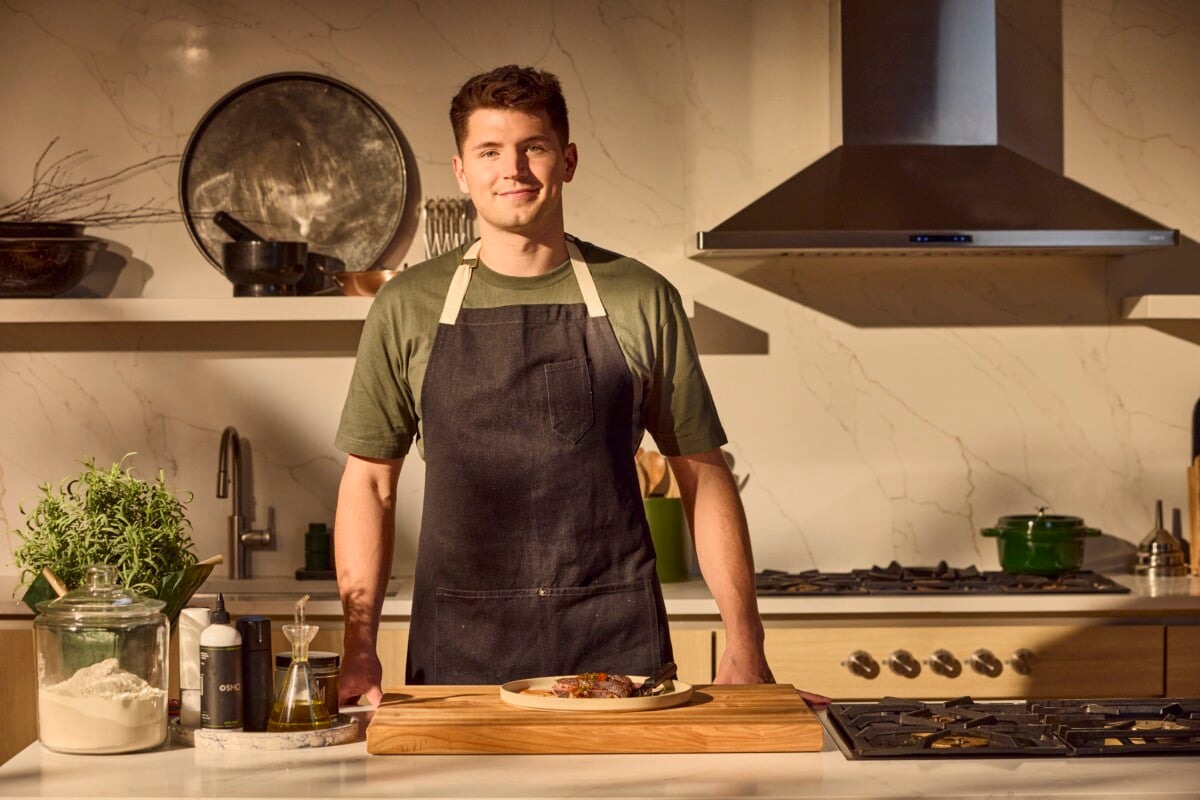 A male chef behind a kitchen counter with a plate of steak in front of him.