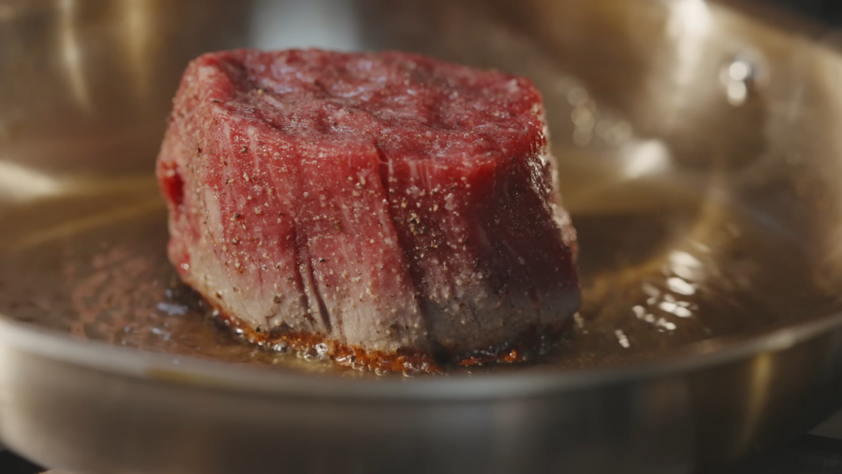 A small steak cooking in a stainless steel pan.