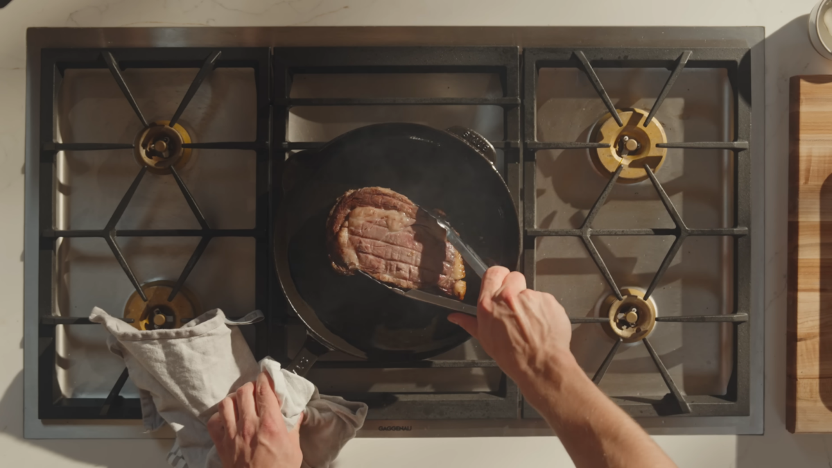 A man flipping a steak with tongs on a cast iron skillet.