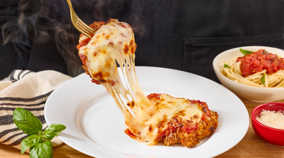 A fork holding a bite of chicken Parmesan above a plate. 