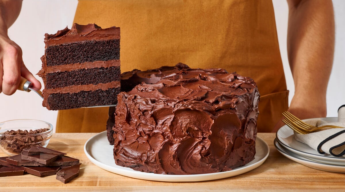 A slice of chocolate layer cake being lifted from a platter with the rest of the cake.