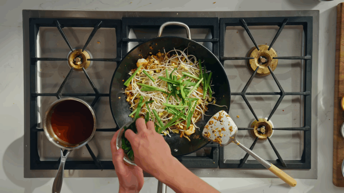 Pad Thai cooking in a wok topped with bean sprouts and garlic chives.