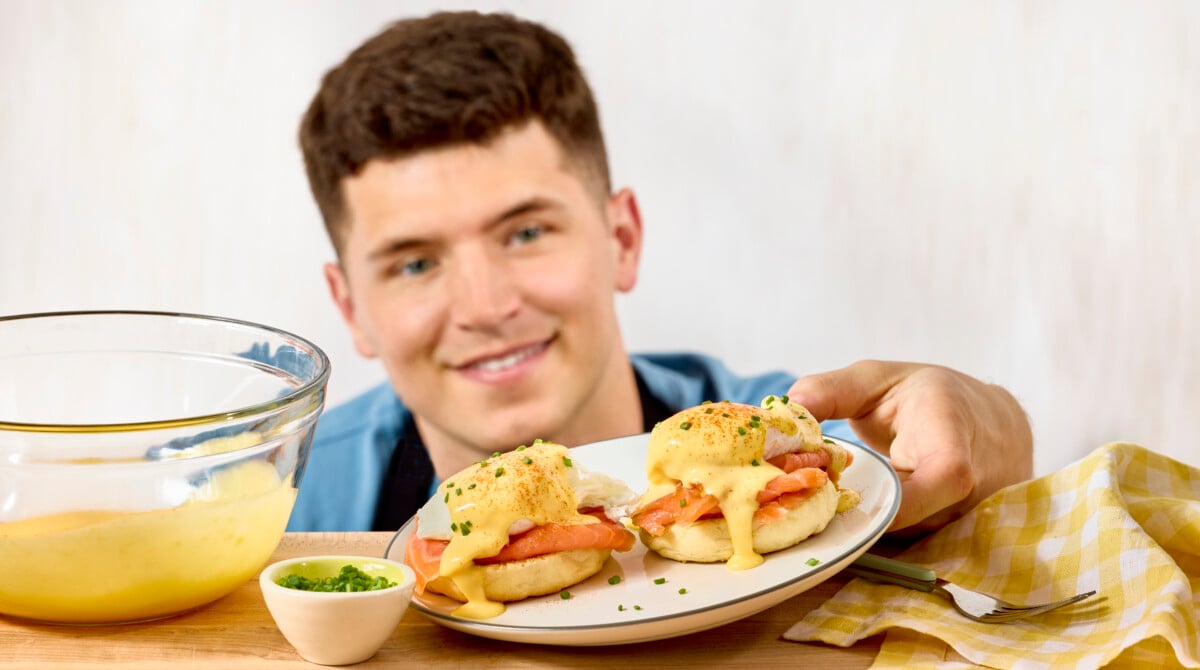 A man kneeling down holding a plate of eggs Benedict made with salmon.