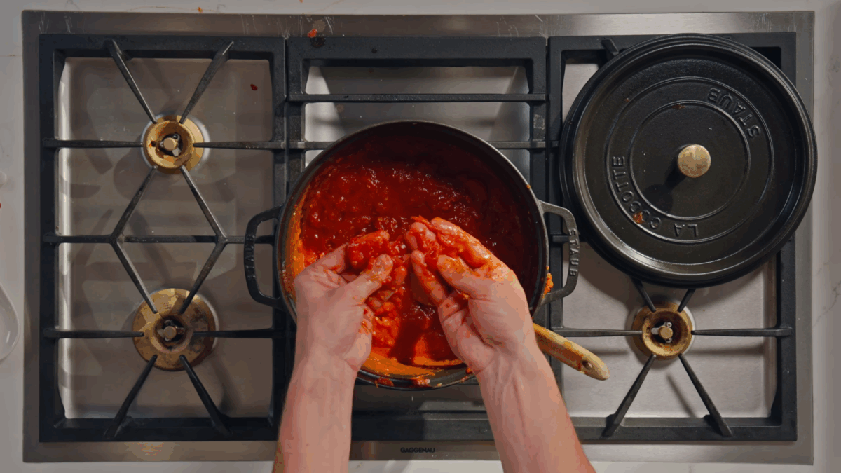 Whole peeled tomatoes being crushed with hand over a Dutch oven.