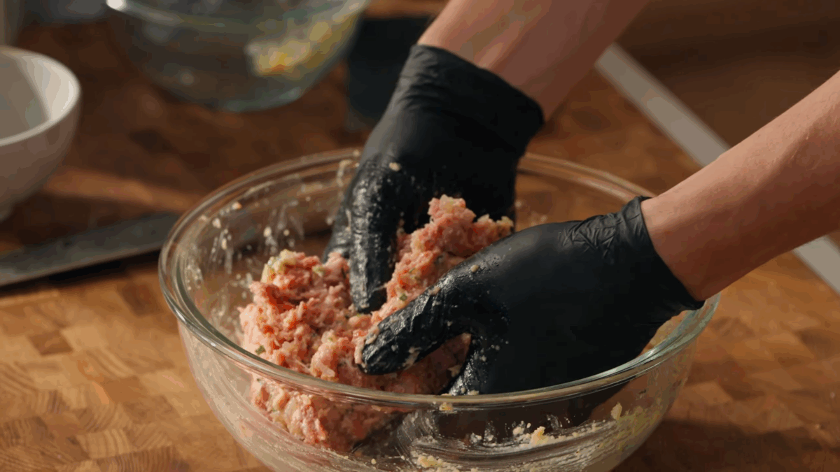A meatball mixture being mixed in a bowl with gloved hands.