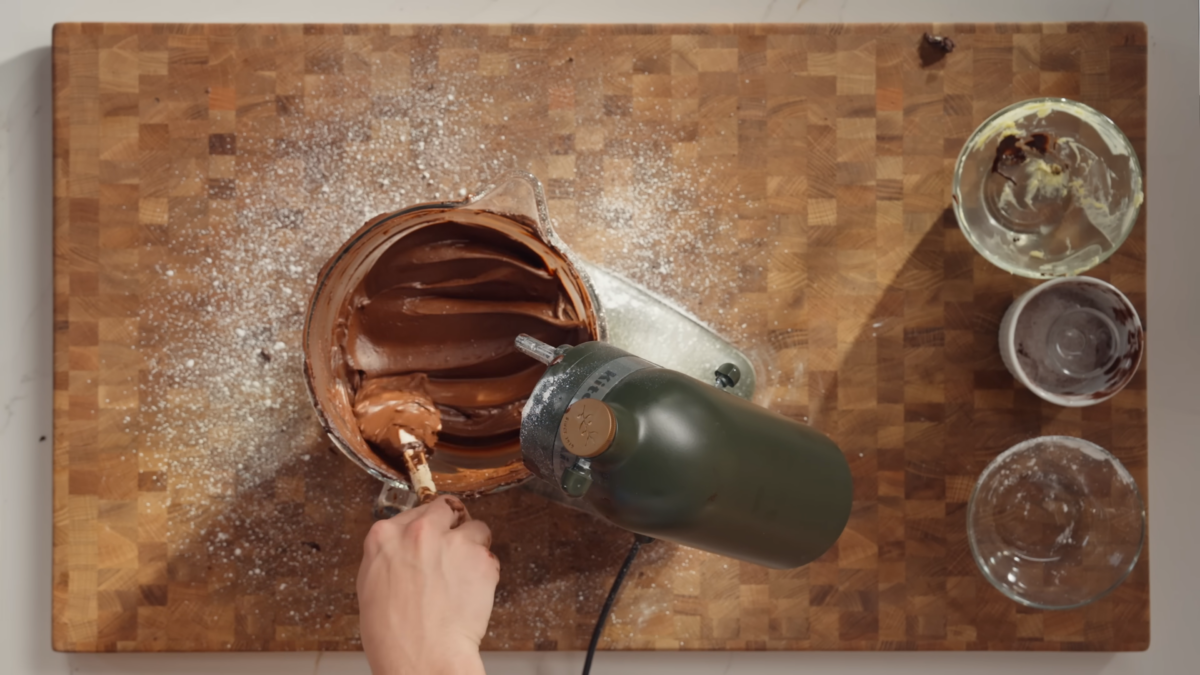 A man mixing chocolate frosting in a glass bowl under an electric mixer.