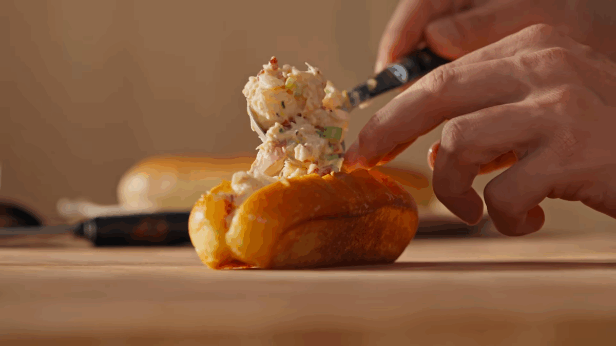 Lobster roll filling being spooned into a toasted New England-style bun.
