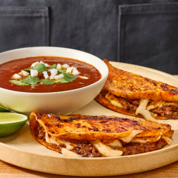 Two beef birria tacos on a plate with a bowl of consomé sauce.
