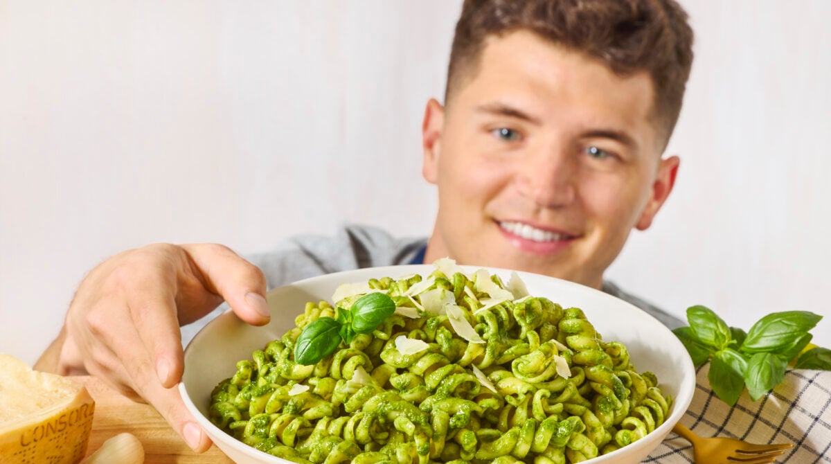 A man grabbing a bowl of basil pesto pasta topped with Parmesan cheese and fresh basil. 