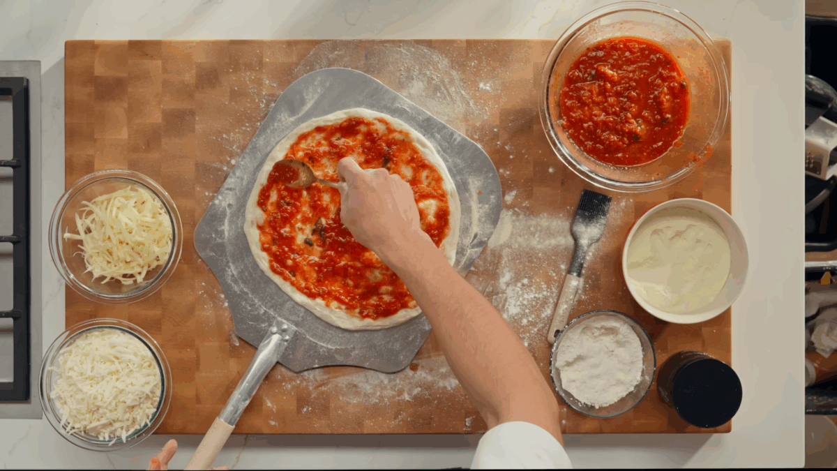 Raw pizza dough being topped with tomato sauce. 