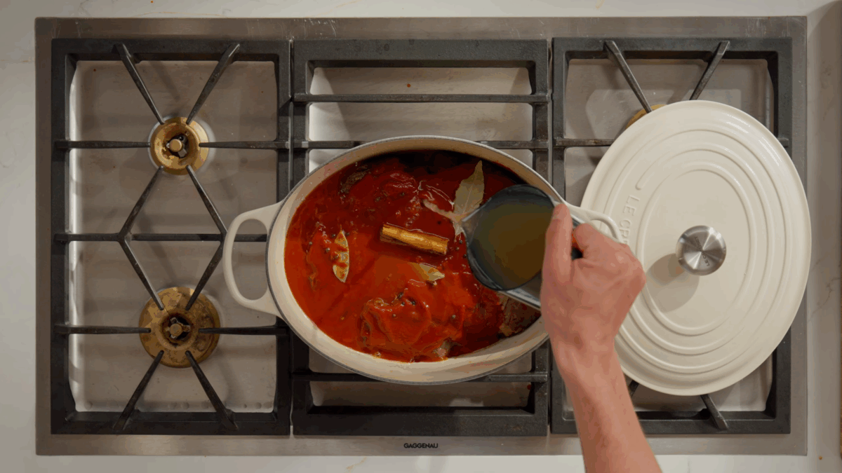 Broth being poured into a pot of beef and a tomato mixture for braising.