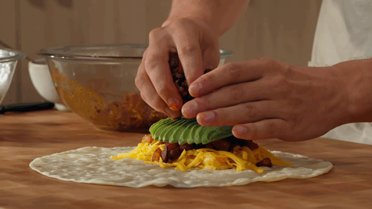 Eggs, sausage, veggies, and avocado being layered on a tortilla. 