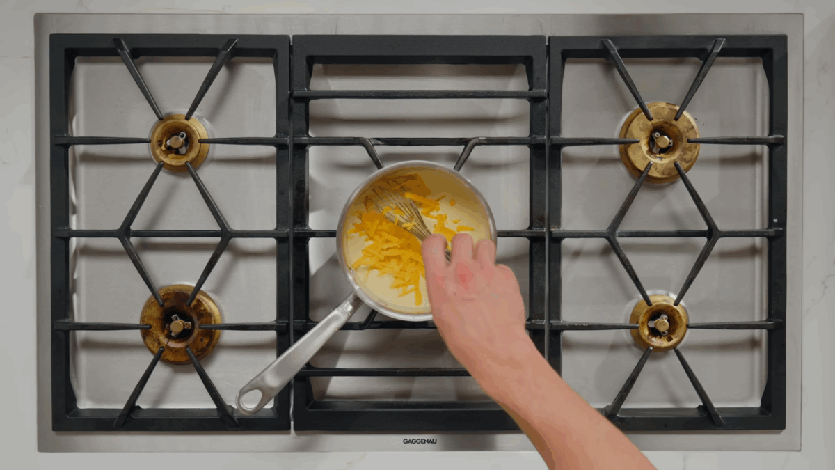 Cheese being added to a milk sauce in a saucepan on a stove. 
