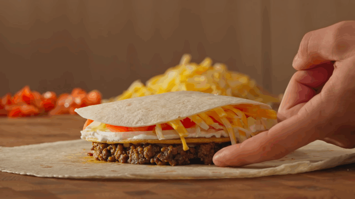 Tortillas, beef, sour cream, tomatoes, lettuce, and cheese being layered to make a crunchwrap. 