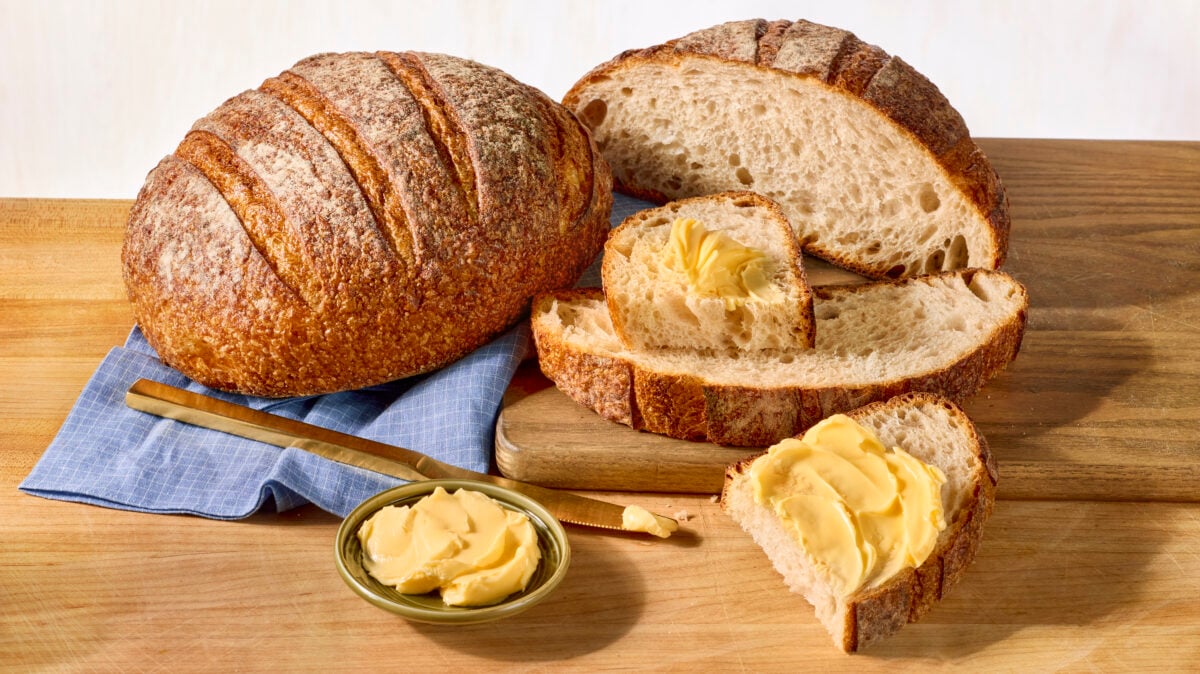 A whole loaf of sourdough bread and a half loaf with two slices cut and two pieces of one slice topped with butter.
