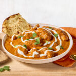 A bowl of butter chicken with a piece of naan bread sticking out.