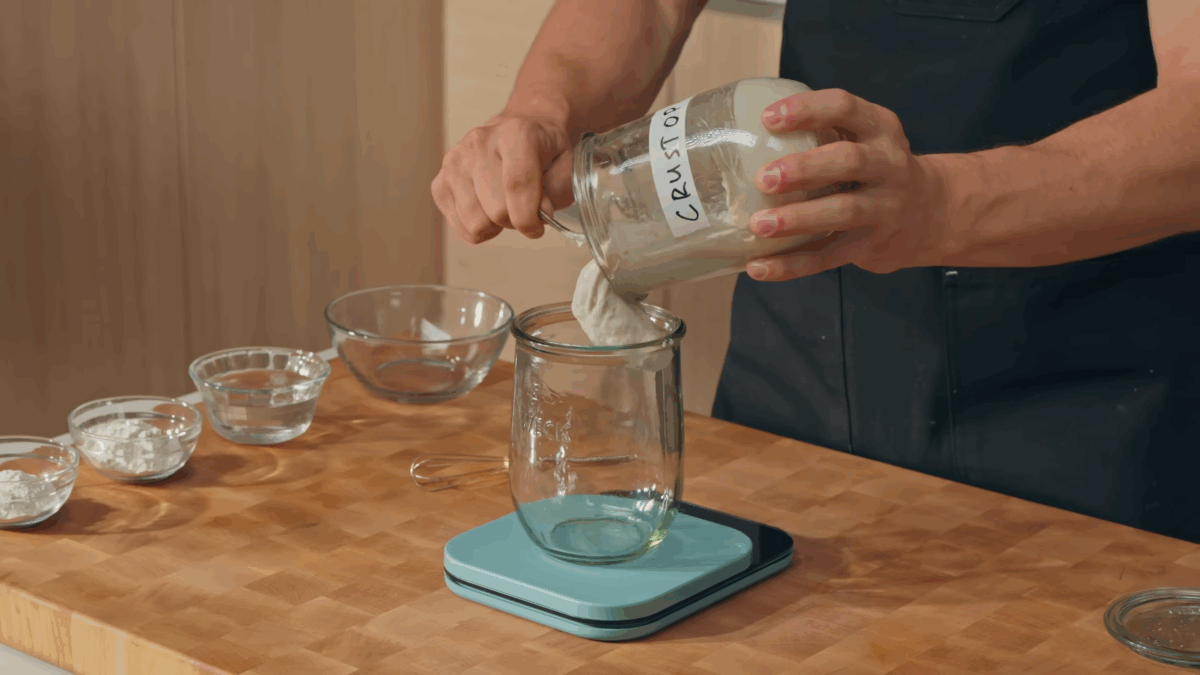 A jar of sourdough starter being poured into another glass jar on a food scale.