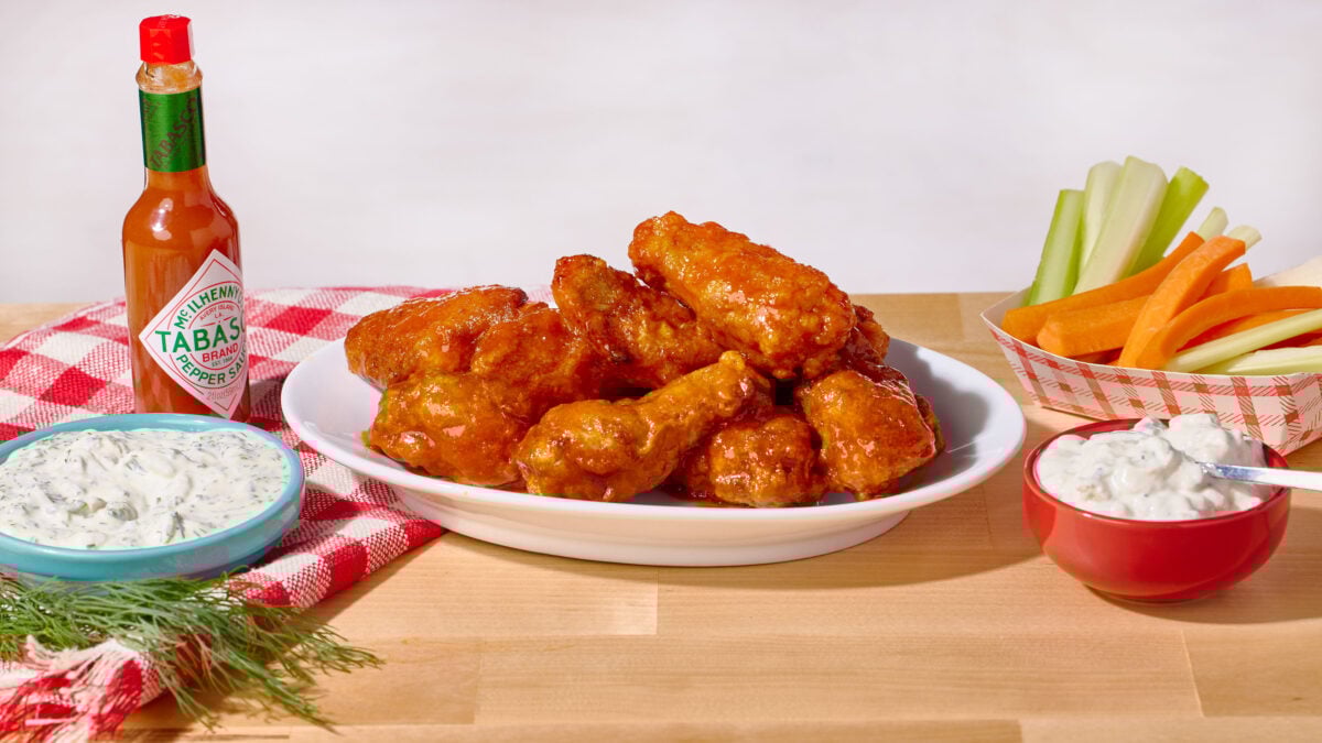 A plate of buffalo chicken wings next to a bowl of blue cheese dressing, a bowl of ranch, a bottle of hot sauce, and celery and carrot sticks.