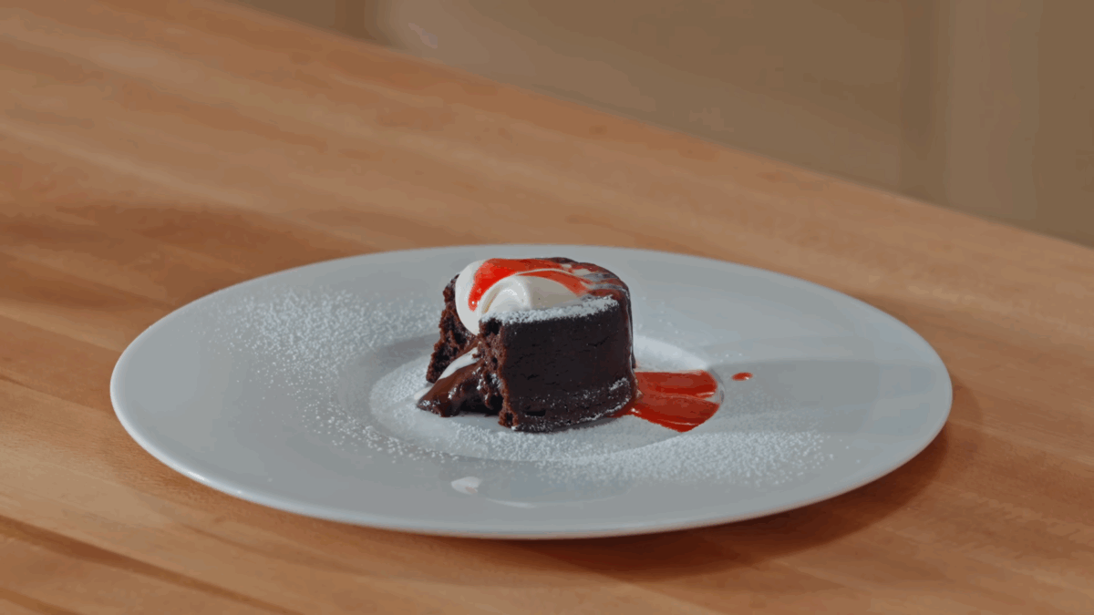 A molted chocolate lava cake on a plate topped with powdered sugar, whipped cream, and raspberry coulis, a bite missing.