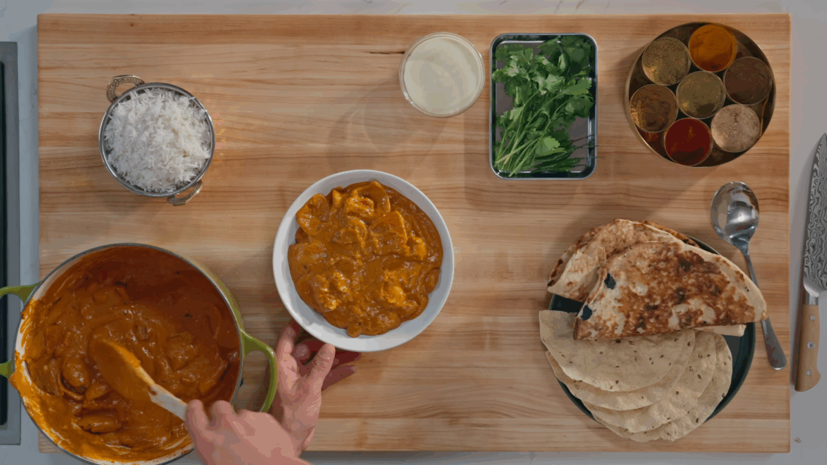 A bowl of chicken tikka masala, basmati rice, naan, pappadum, and fresh herbs.