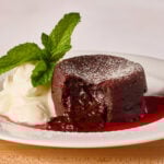 A molten chocolate lava cake on a plate with raspberry coulis, whipped cream, and a mint leaf, a bite missing from the cake.